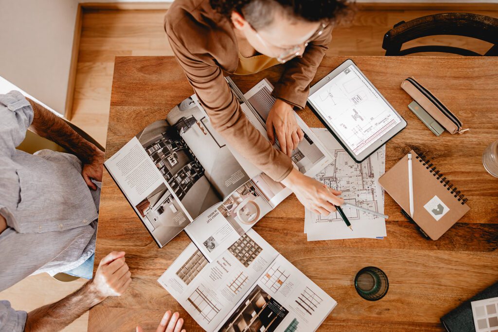Eine Immobilienberaterin bespricht mit ihrem Kunden innovative Gestaltungs- und Umbaukonzepte für eine Wohnung anhand von Plänen und Katalogen am Tisch.