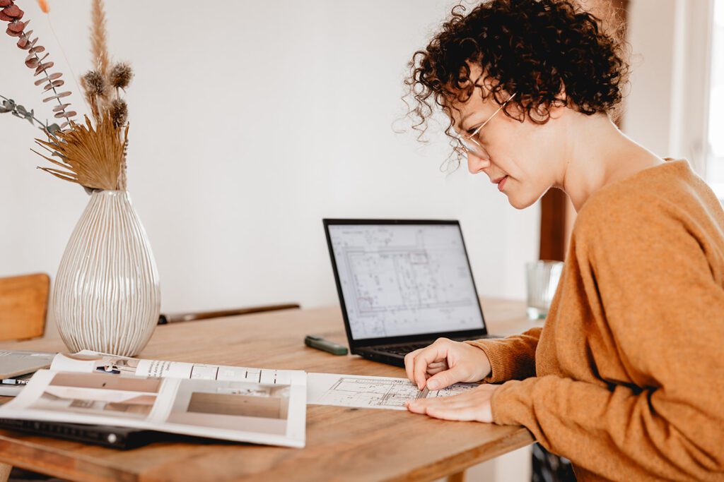 Immobilienprofi sitzt am Arbeitsplatz, sichtet Verkaufsunterlagen und erarbeitet die Grundrissplanung einer Immobilie.