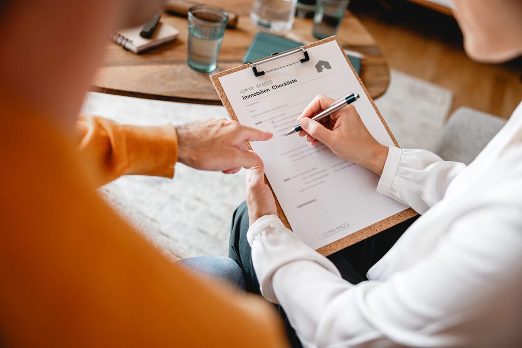 Eine Immobilienberaterin bespricht gemeinsam mit einem Kunden bei einer Wohnungsbesichtigung eine Checkliste für den Wohnungskauf.